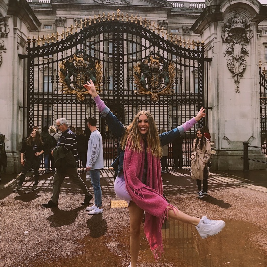 Photo of Candace wearing a pink scarf, purple mini dress, and white Fila tennis shoes from Urban Outfitters, and a blue denim Levi's jacket in front of Buckingham Palace in London, England.
