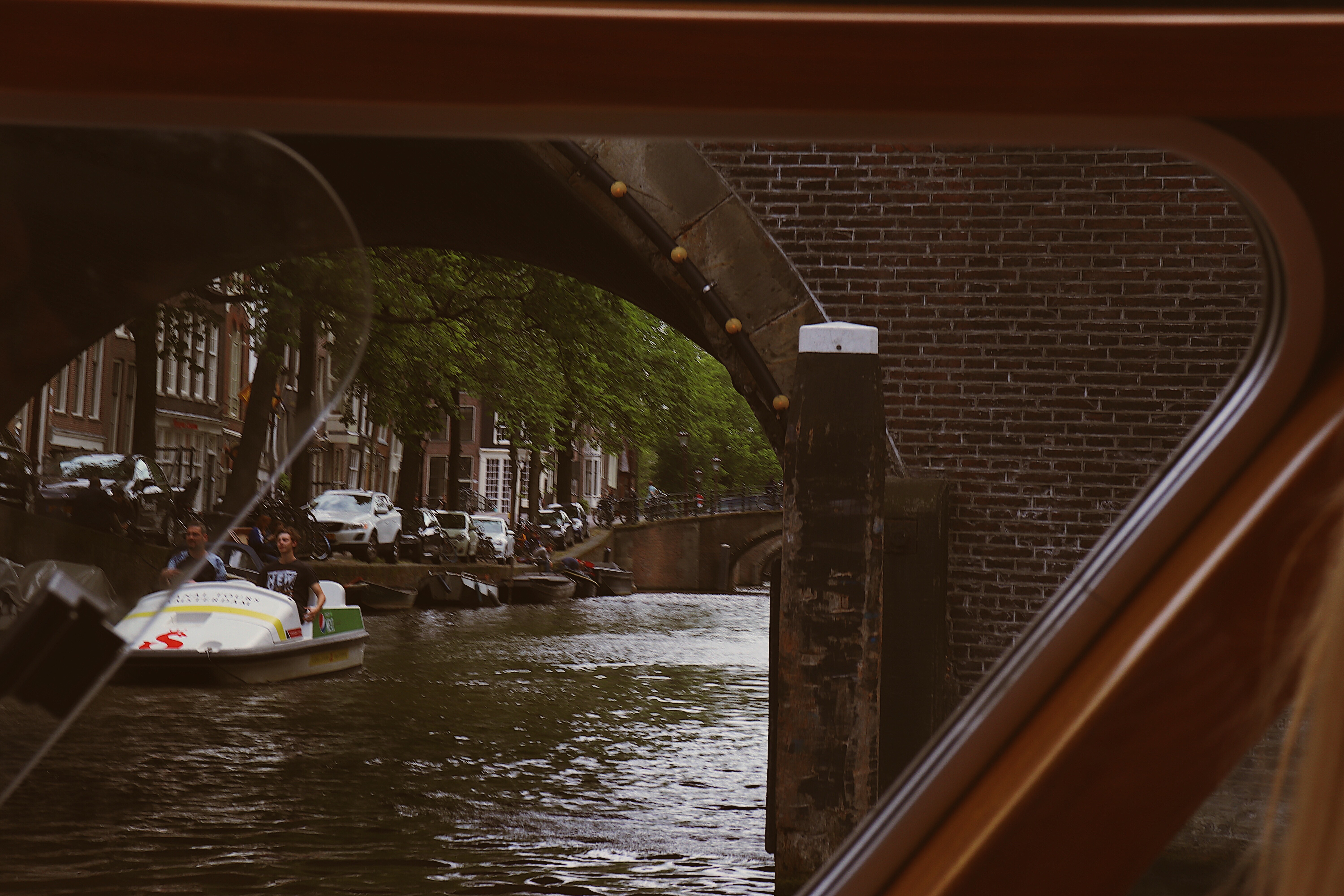 Photo from inside the canal tour boat of people enjoying a day on the canals in a smaller boat from the other side of the bridge in Amsterdam, Netherlands. 
