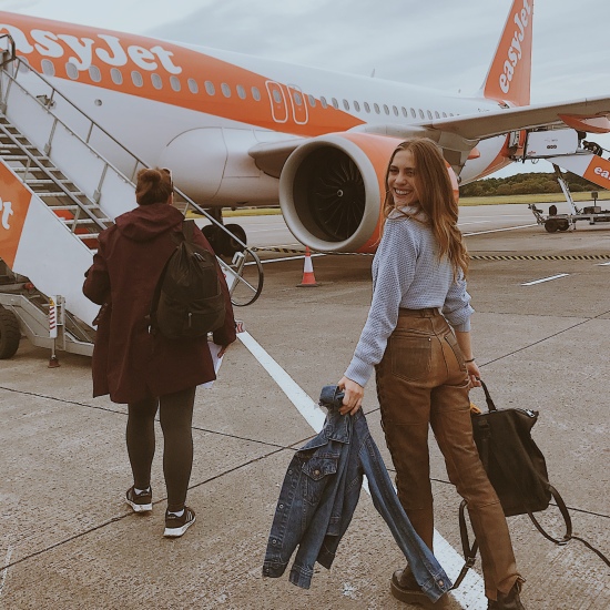Photo of Candace wearing a blue sweater, brown leather pants, and black platform Doctor Martens; holding a black Michael Kors backpack and blue denim Levi's jacket. She is boarding an Easy Jet airplane in Scotland.