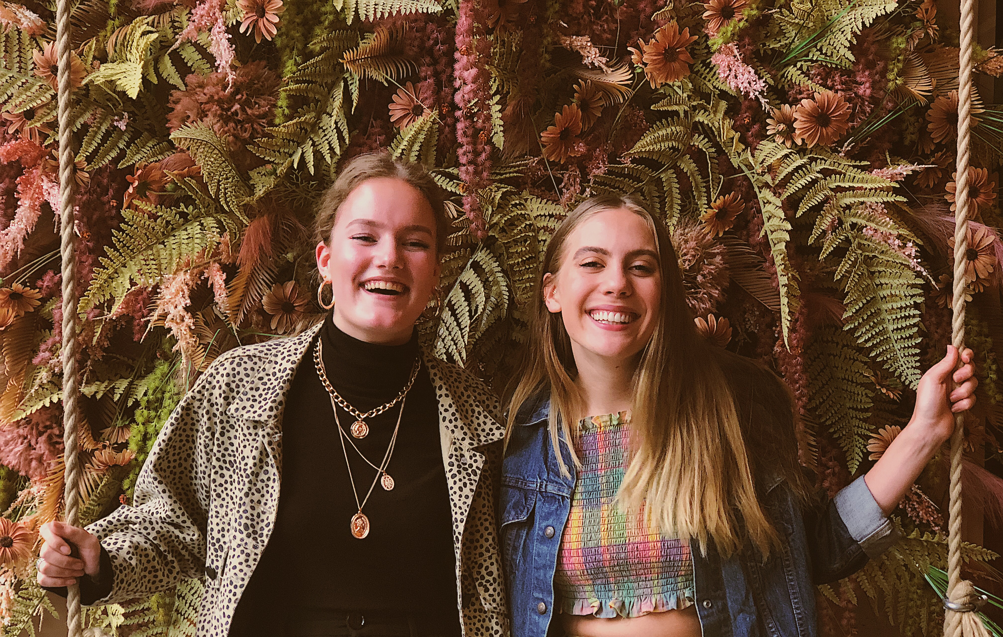 Candace and Mandy at a cafe in front of a flower wall in Amsterdam, Netherlands; Mandy is wearing a black turtle neck, cheetah print jacket, and gold necklaces, Candace is wearing a denim Levi's jacket and plaid crop top..
