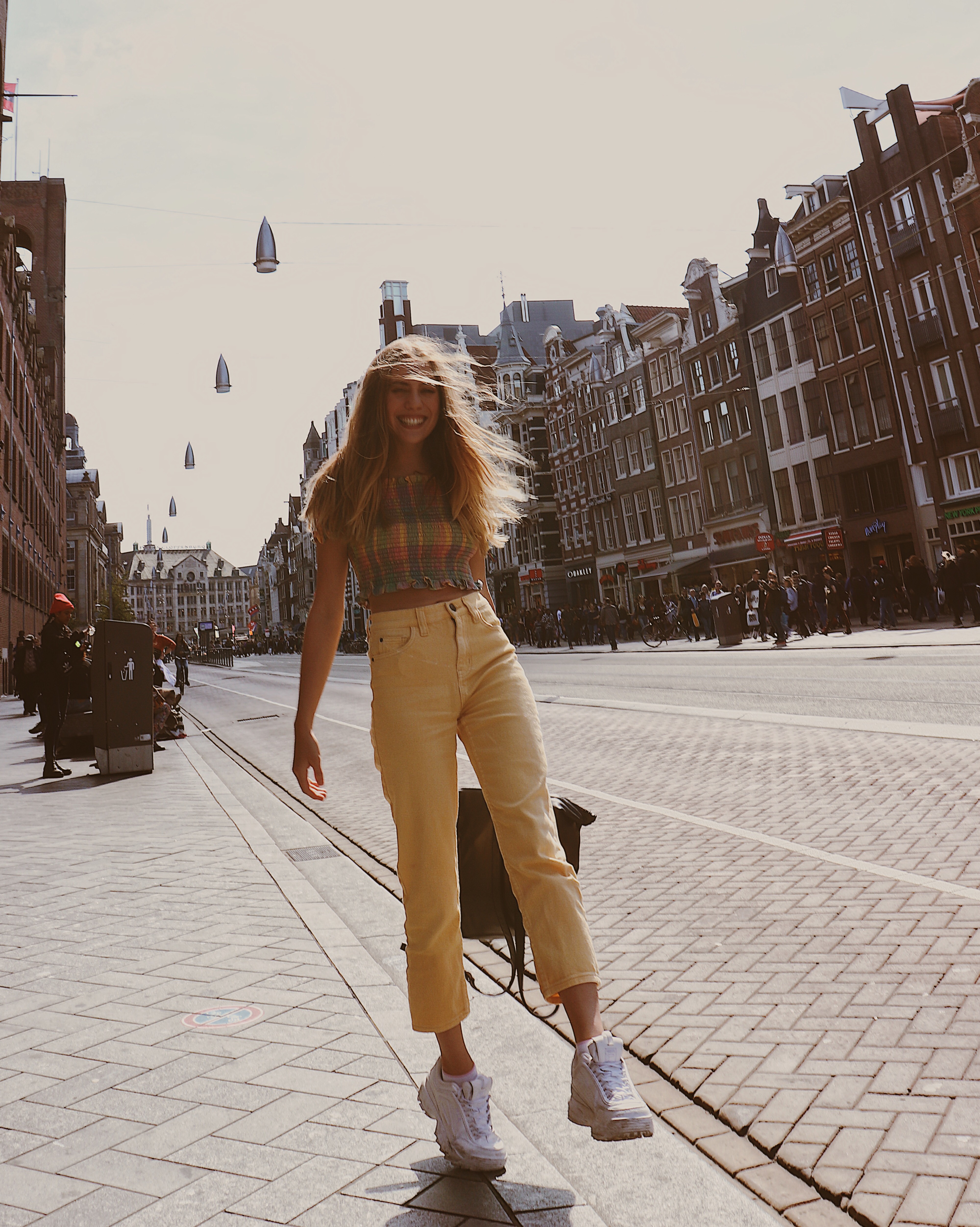 Candace staning on a brick sidewalk in Amsterdam, Netherlands wearing yellow jeans and a pastel plaid crop top form Urban Outfitters and white Fila tennis shoes.