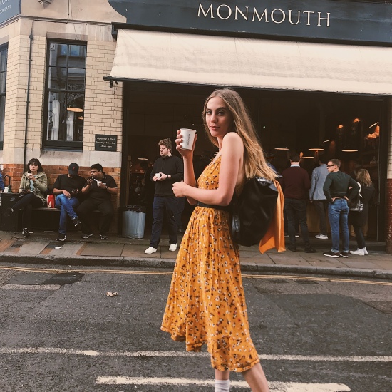 Photo of Candace in a yellow sundress and black slides from Urban Outfitters, white ankle socks from Forever 21, and a black Michael Kors backpack in front of Monmouth Coffee in Borough Market in London England holding a cup of coffee.