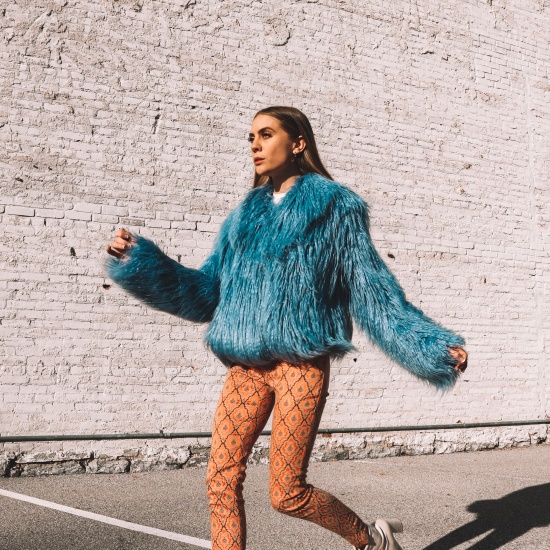Candace in a blue faux fur jacket, orange and brown printed skinny pants, and cream boots in front of a brick wall during golden hour.