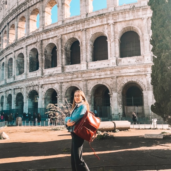 Candace posing in front of the Colosseum in Rome, Italy with her red Italian leather backpack , wearings a Levi's denim jacket, Urban Outfitters black jeans, grey turtleneck, and black Doc Martens.