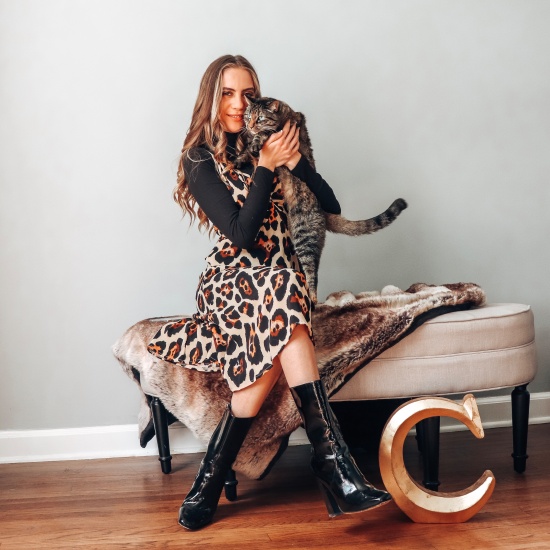 Candace sitting and smiling in a leopard dress from Nasty Gal, black turtleneck, and black shiny calf boots with her cat Fifi.