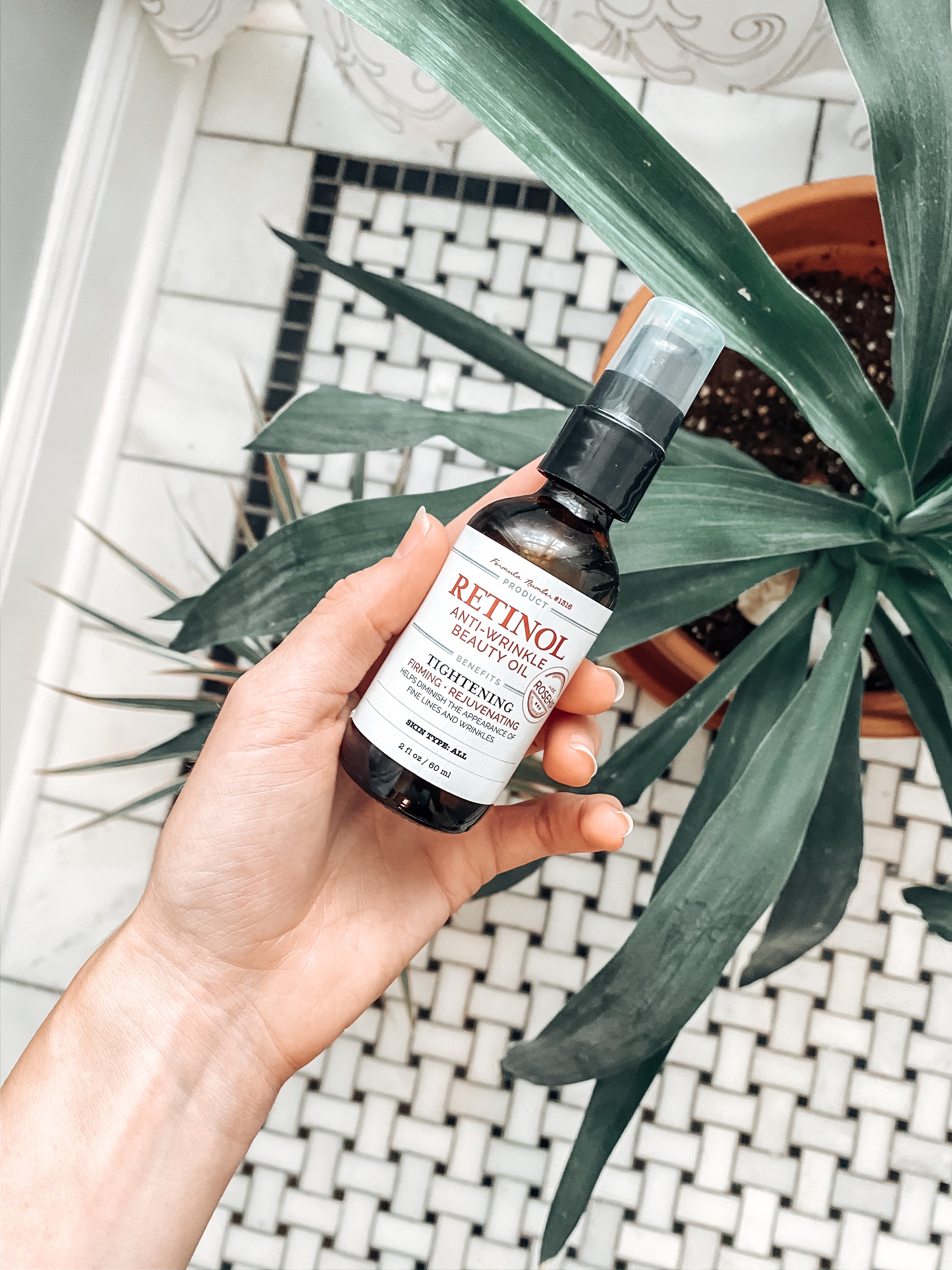 Image of Candace's hand holding a bottle of Retinol Oil over plants and black and white marble tile.