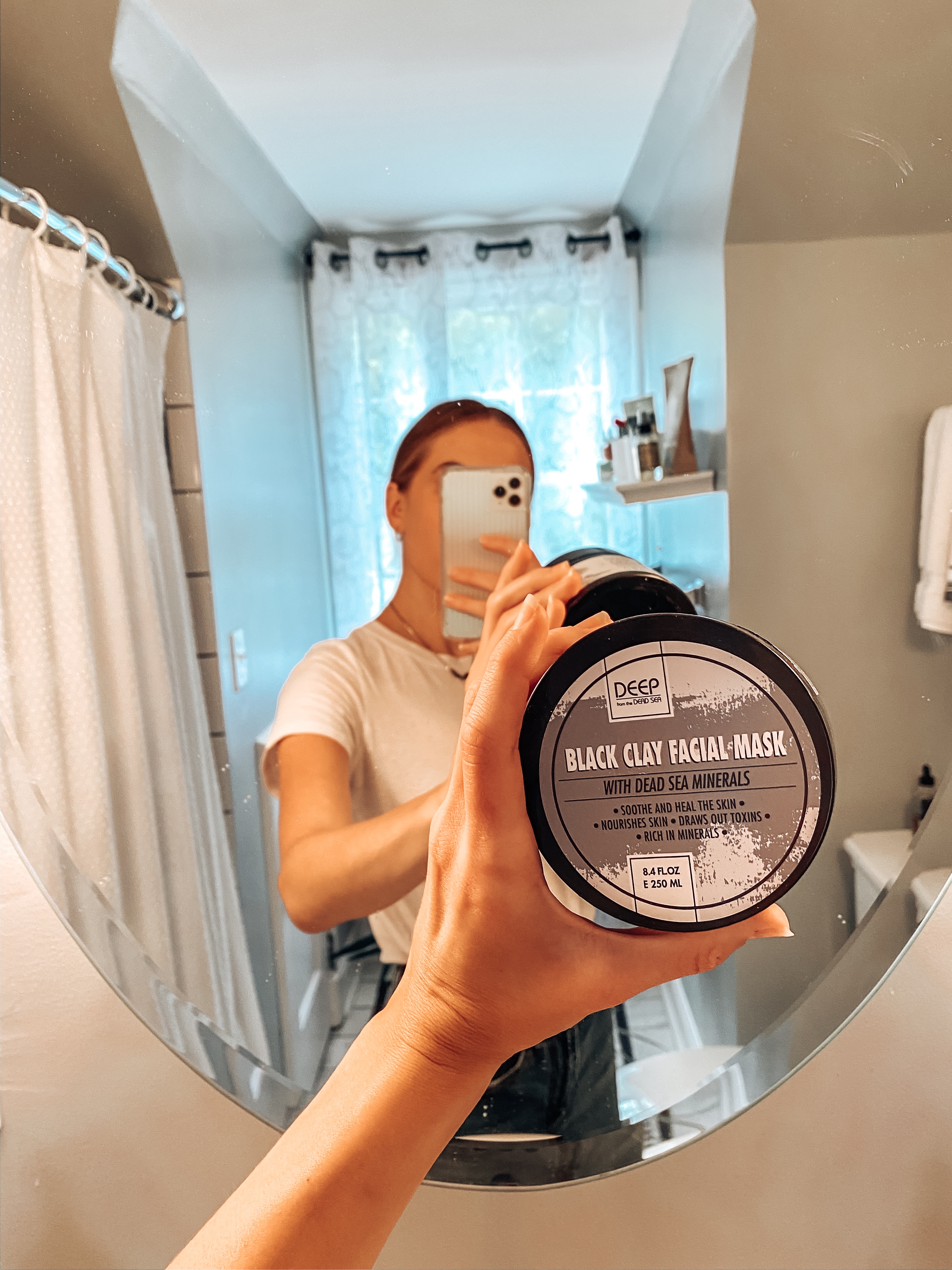 Image of Candace holding up a Black Clay Facial Mask in front of a mirror in her bathroom. 